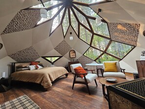 Ein Zimmer mit einem geometrischen Deckendesign, einem Bett, einem Sessel, einem Tisch und Blick auf Bäume durch große Fenster.