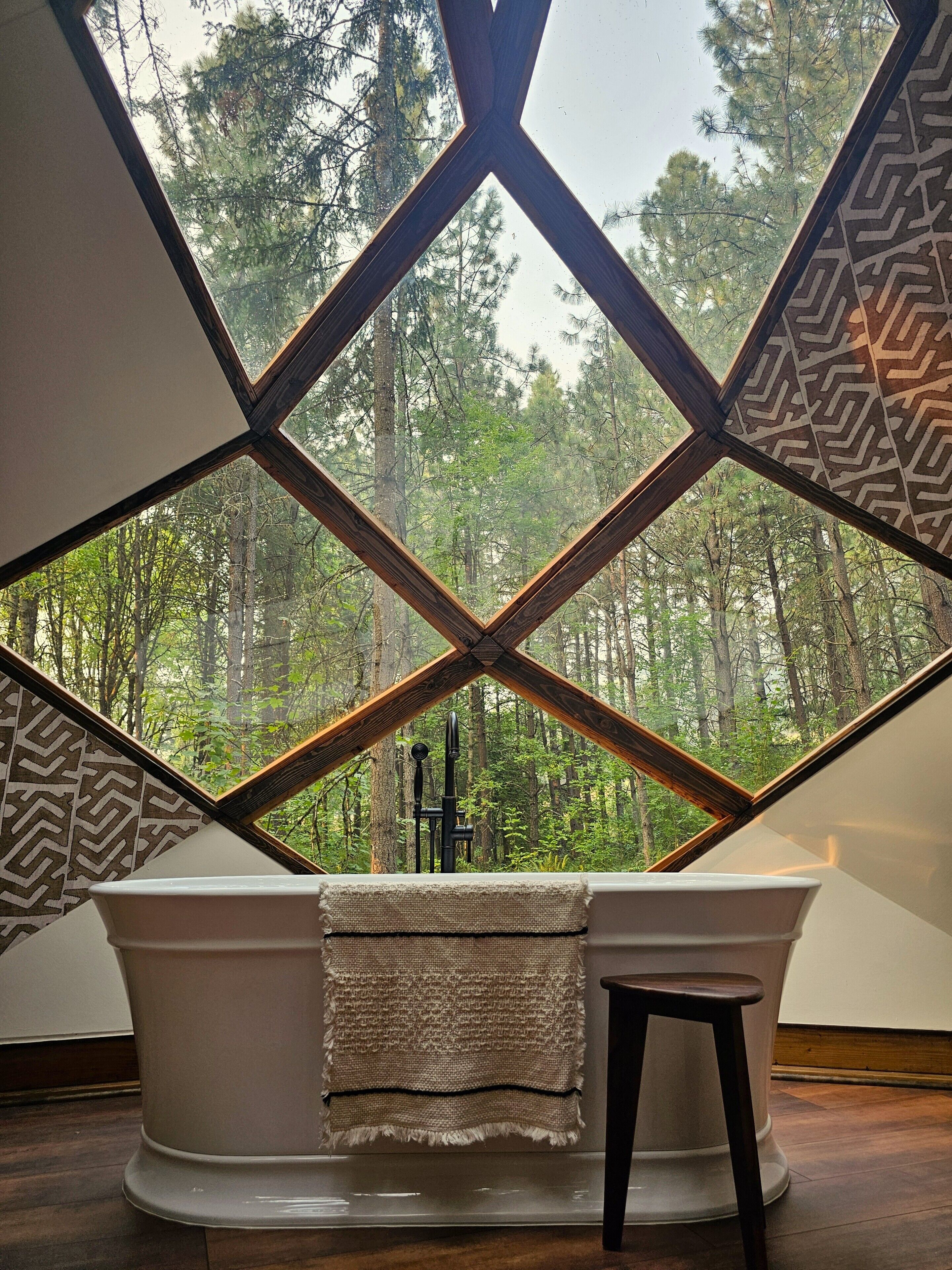 Une baignoire offrant une vue sur une forêt, grâce à une structure de fenêtre aux motifs géométriques.