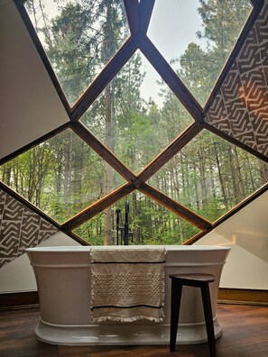 Eine Badewanne mit Blick auf einen Wald, der durch eine geometrische Fensterkonstruktion zu sehen ist.