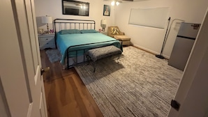 A bedroom with a bed, a nightstand, a chair, a ceiling fan, a window with blinds, and a refrigerator.