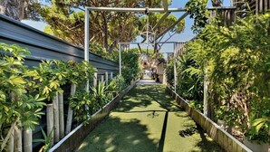Un étroit sentier bordé d’une végétation luxuriante et d’une clôture en bois, qui mène à un bâtiment doté d’un toit en pente.