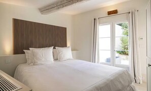 Une chambre à coucher avec un lit, du linge de lit blanc, une tête de lit à lattes et une fenêtre avec des rideaux.