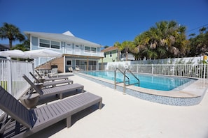A pool area with lounge chairs and a building in the background. - The Woods (Fort Myers Beach)