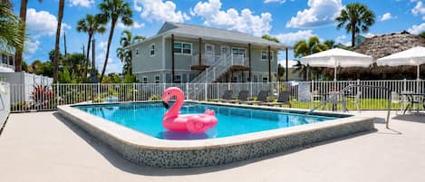 A swimming pool with a pink flamingo float, surrounded by a white fence, palm trees, and a building with a balcony.