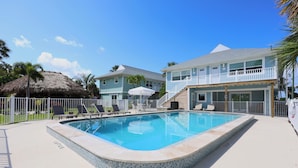 Ein Swimmingpool mit Fliesenrand, Liegestühlen und einer strohgedeckten Struktur.