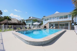 Ein Swimmingpool mit Fliesenrand, Liegestühlen und einer strohgedeckten Struktur.