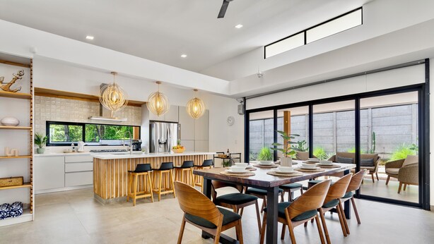 A modern kitchen with a central island, dining area with wooden chairs, and large windows offering a view of greenery. - Villa con Piscina Privada a 3 min de la Playa / Concierge incluido (Playa Grande)