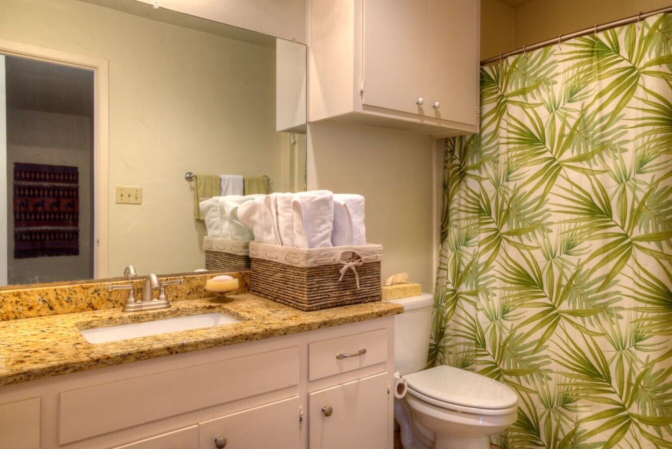 Baño con encimera de granito, espejo, lavabo, inodoro y cortina de ducha con estampado de hojas.