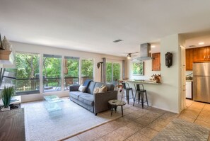 A modern living room with a grey sofa, a coffee table, and a dining area with bar stools. Large windows provide a view of the outdoors. - 5 bed/5 bath Hiking on Greenbelt Close to Zilker! (Austin)