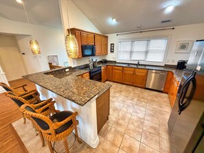 A kitchen with granite countertops, wooden cabinets, a stainless steel refrigerator, and a built-in island with bar stools. - Serenity Sound House Sandbridge Oasis With Pool (Virginia Beach)