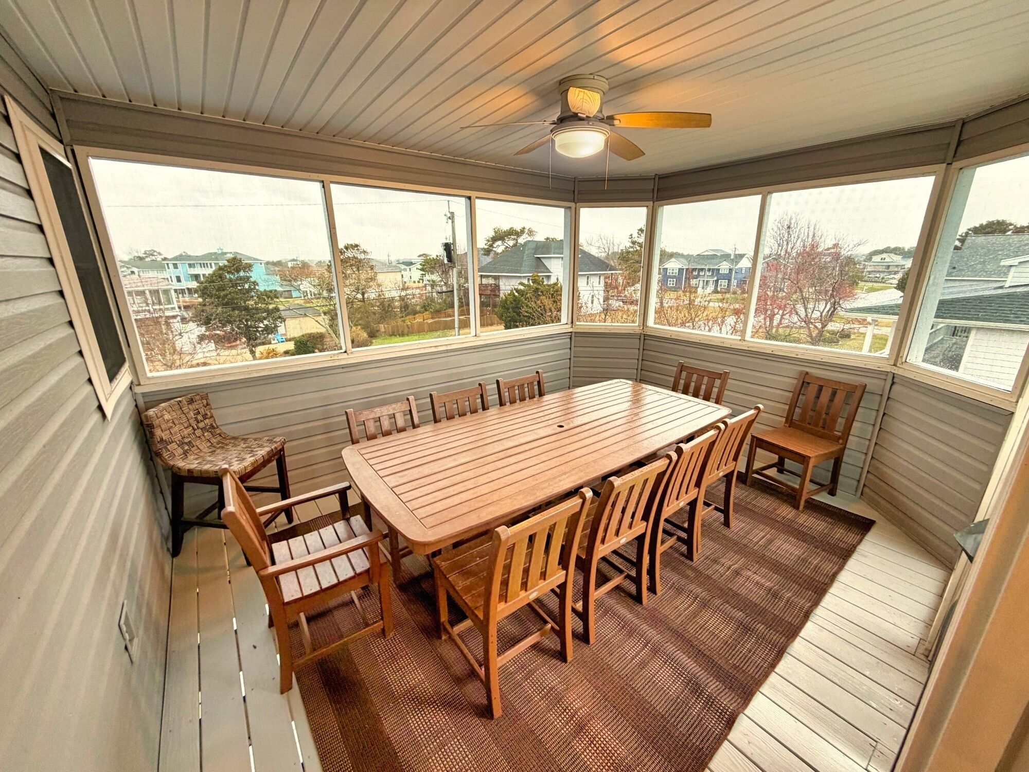 A covered patio with a wooden table and chairs, large windows, and a ceiling fan.