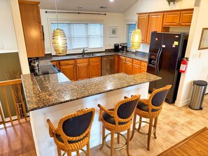 A kitchen with a granite countertop, wooden cabinets, a stainless steel refrigerator, and three wicker bar stools. - Serenity Sound House Sandbridge Oasis With Pool (Virginia Beach)