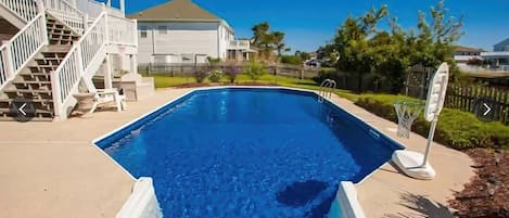 A swimming pool with a clear blue water surface, surrounded by a concrete deck, a white fence, and a staircase leading to a building.