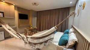 Ein modernes Hotelzimmer mit einer Hängematte, einem Bett mit gemusterten Betttextilien, einem kleinen Tisch mit Stühlen, einem Fernseher und einer Küchenzeile.