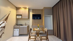 Ein kompaktes Hotelzimmer mit Küchenzeile, einem Essbereich mit zwei Stühlen, einem flachbildfernseher an der Wand und einem großen Fenster mit Vorhängen.