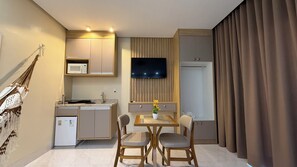 Een compacte hotelkamer met kitchenette, een zithoek met twee stoelen, een flatscreen televisie aan de muur en een groot raam met gordijnen.