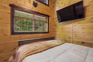 A wooden cabin room with a bed, a window with blinds, and a flat-screen TV mounted on the wall. - Brand New!  2-bedroom cabin with AC in scenic Sevierville. (Sevierville)
