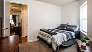 A bedroom with a bed, a desk, a window, and a door leading to another room.