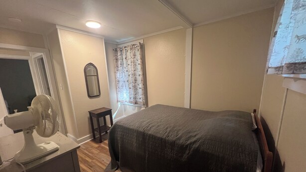Un dormitorio con cama, ventilador, una mesita, espejo y ventana con cortinas.