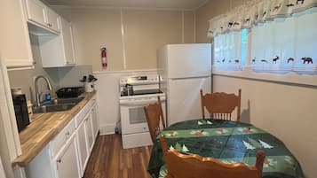 Une cuisine compacte avec des meubles blancs, un four à micro-ondes, une cuisinière et un réfrigérateur. On y trouve une table à manger avec des chaises et une fenêtre agrémentée de rideaux à motifs animaliers.
