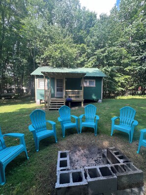A small cabin with a porch and a fire pit surrounded by four chairs.