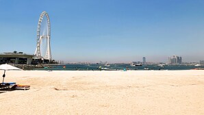Ein Strand mit einem großen Riesenrad im Hintergrund, einem sandigen Ufer und mehreren Booten auf dem Wasser.