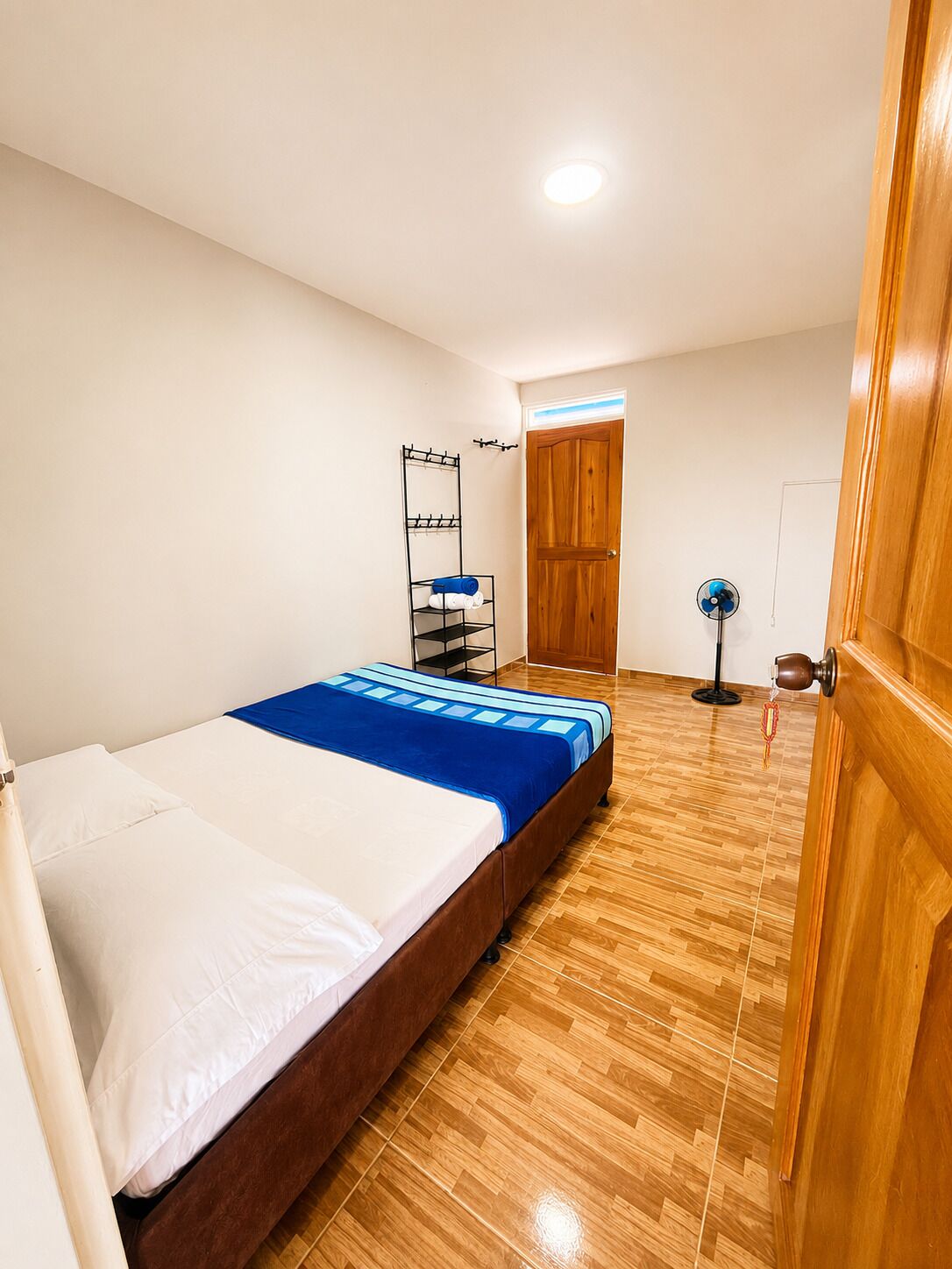 A bedroom with a bed, wooden flooring, a wardrobe, a fan, and a wall-mounted shelf.