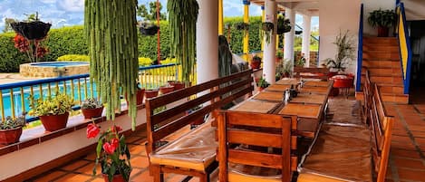 Ein überdachter Außenbereich mit Holzbänken und -tischen, Topfpflanzen und Blick auf einen Pool und viel Grün.