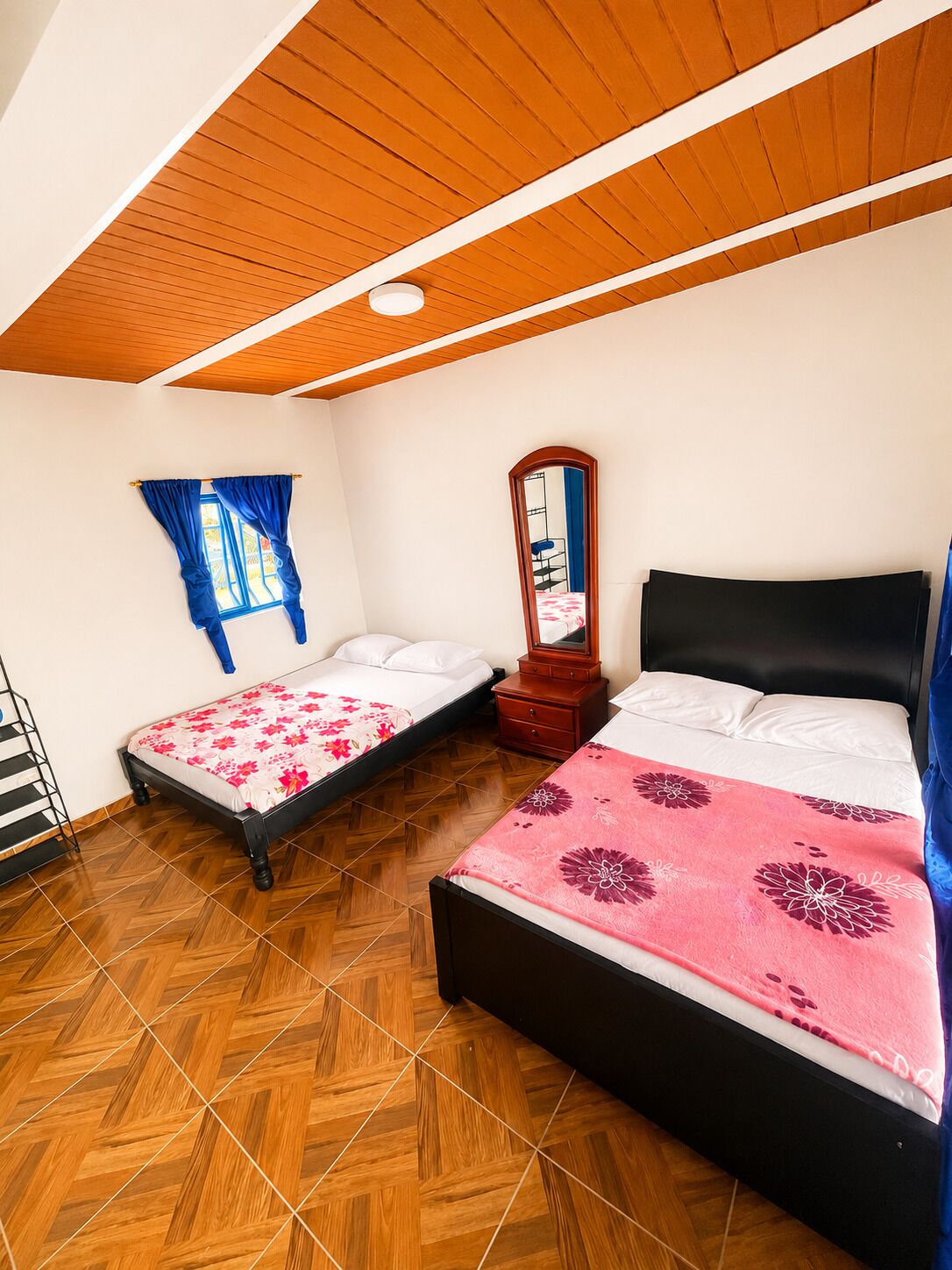 A bedroom with two beds, a wooden ceiling, a window with blue curtains, and a mirror.