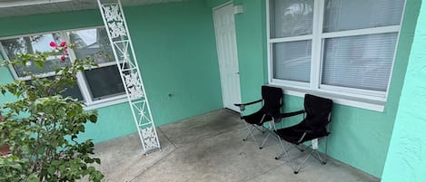 A covered outdoor area with a concrete floor, two black chairs, a white ladder, and a green wall with windows.