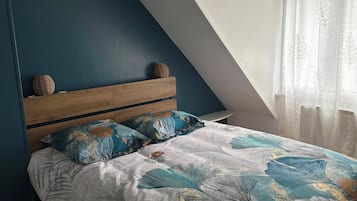 Et soveværelse med skråvæg, træsenggavl, blomstret dyne og et vindue med lette gardiner.