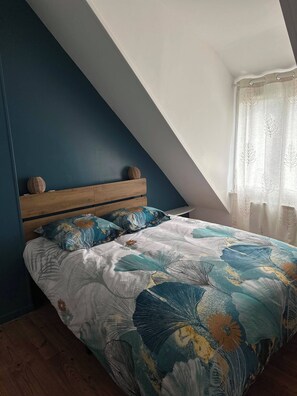 A bedroom with a sloped ceiling, a wooden headboard, a floral bedspread, and a window with sheer curtains. - Just 5 minutes from the harbor and 1,500 meters from the beaches, this comfortable home is perfect for a relaxing stay! (Douarnenez)