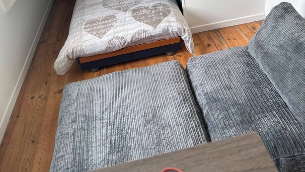 A small bedroom with a slanted ceiling, a single bed with patterned bedding, a nightstand with a lamp, and a grey sofa.