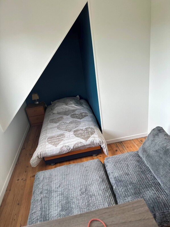 A small bedroom with a slanted ceiling, a single bed with patterned bedding, a nightstand with a lamp, and a grey sofa.