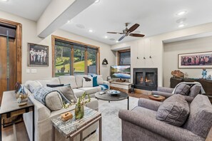 A modern living room with a fireplace, a ceiling fan, and large windows. - Fairway Escape | Private Hot Tub & Views (Avon)