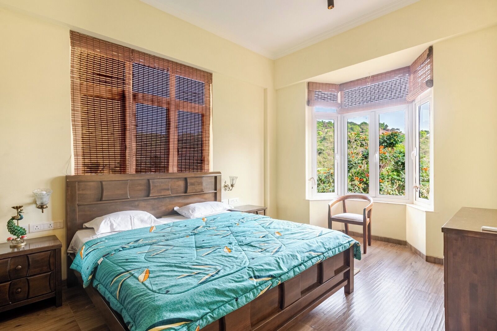 Un dormitorio con cama, una silla, una mesita de noche, una lámpara y una ventana que ofrece vistas al verdor.