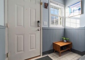 Ein Flur mit einer weißen Tür, einer hölzernen Bank mit einer Pflanze und einem Fenster mit Jalousien.