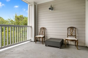 A balcony with two chairs and a small table, a light fixture on the wall, and a view of greenery. - Furnished Monthly Rental near Folly (Charleston)