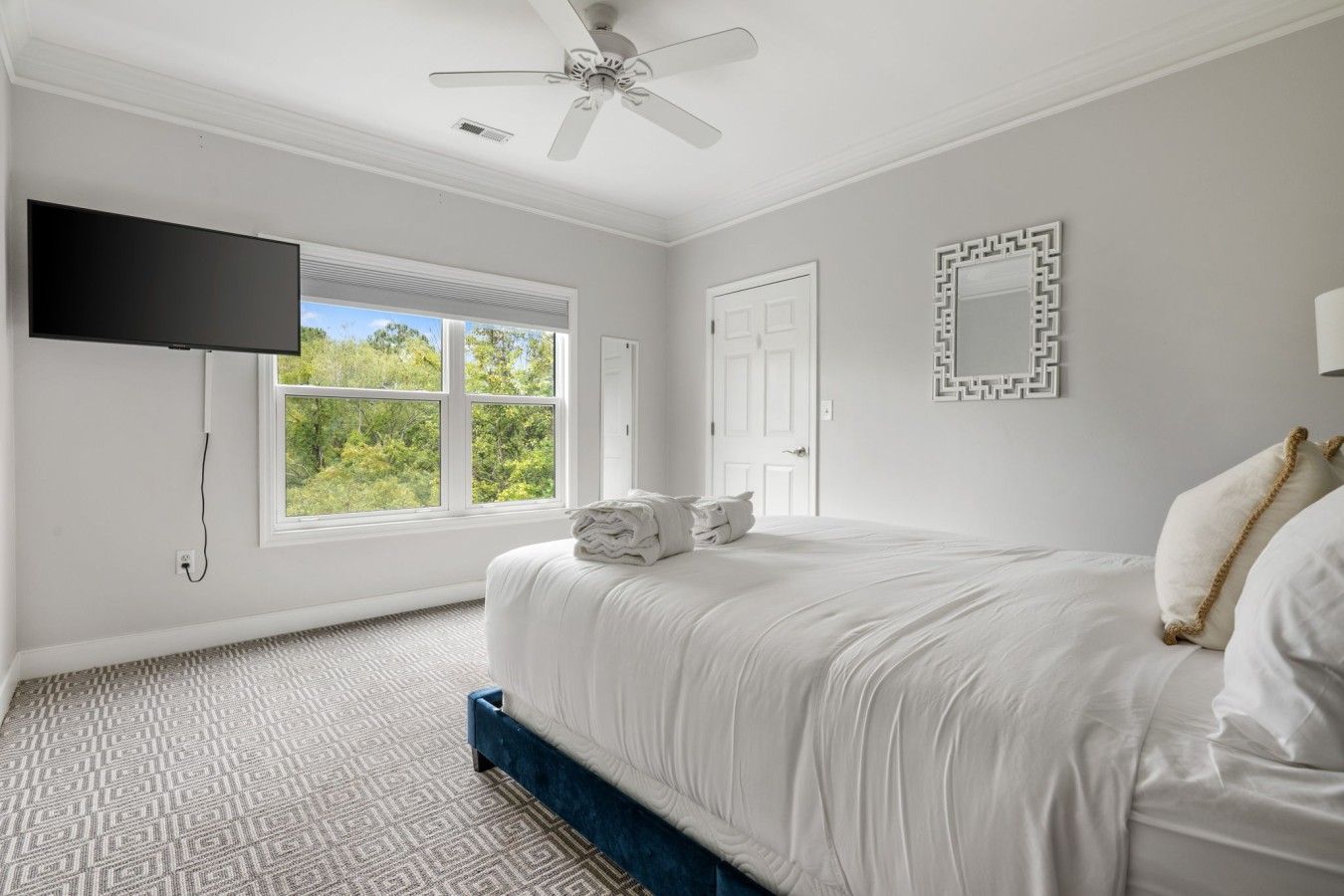 Un dormitorio con cama, ventilador de techo, televisor montado en la pared, ventana con vista a follaje y alfombra estampada.