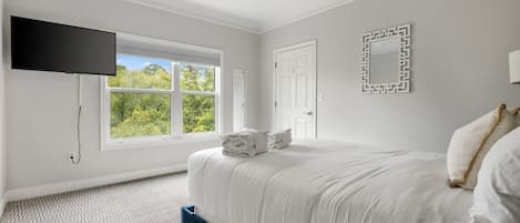 Un dormitorio con cama, ventilador de techo, televisor montado en la pared, ventana con vista a follaje y alfombra estampada.