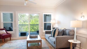 A living room with a sofa, a coffee table, a chair, and a lamp. There are windows with a view of the outdoors. - Furnished Monthly Rental near Folly (Charleston)