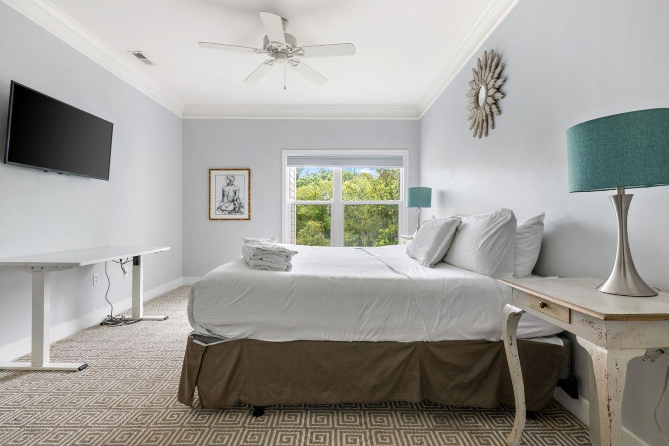 A bedroom with a bed, a desk, a television, a window with a view, and a ceiling fan.