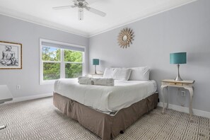 Een slaapkamer met een groot bed, twee bedlampjes, een plafondventilator, een raam met uitzicht op groen en een decoratief object aan de muur.
