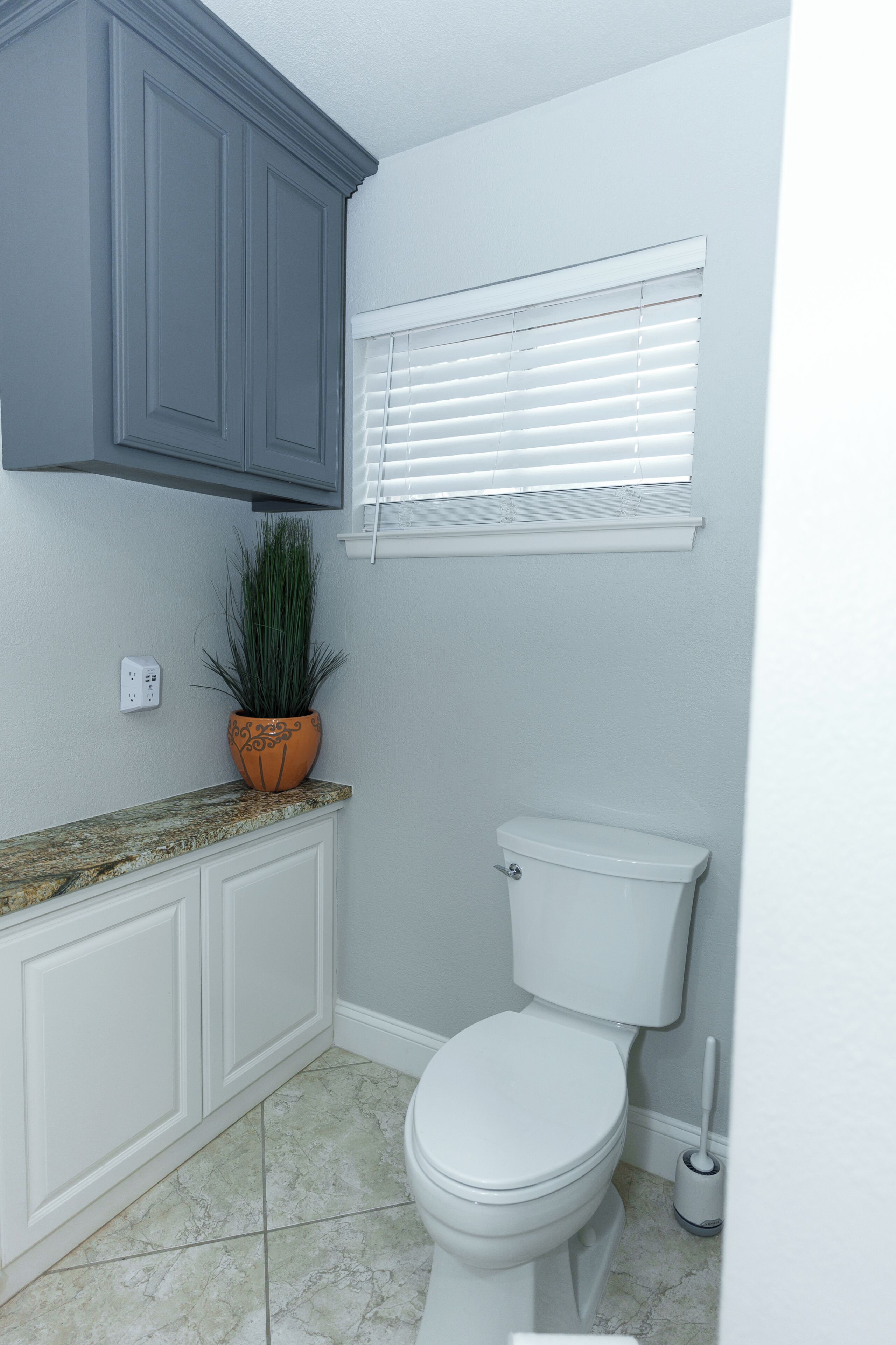 A bathroom with a toilet, a potted plant, a window with blinds, and a granite countertop.