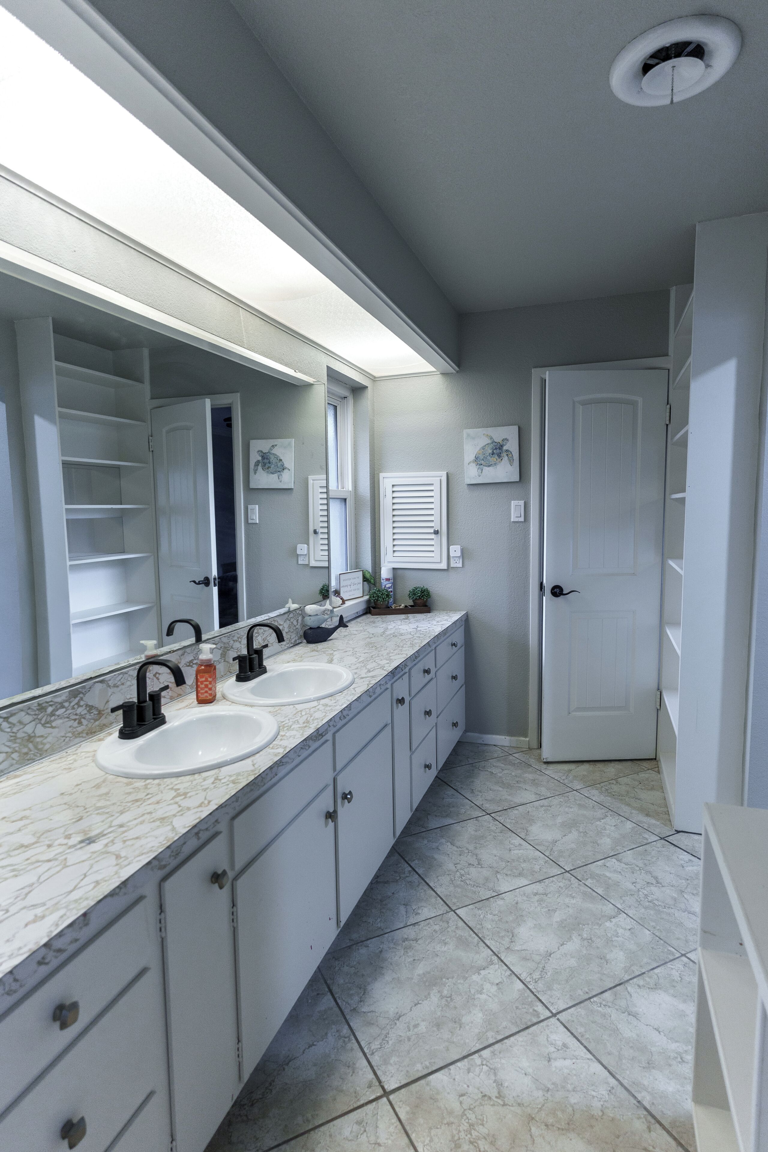 A bathroom with a double sink vanity, a large mirror, and a door leading to another room.