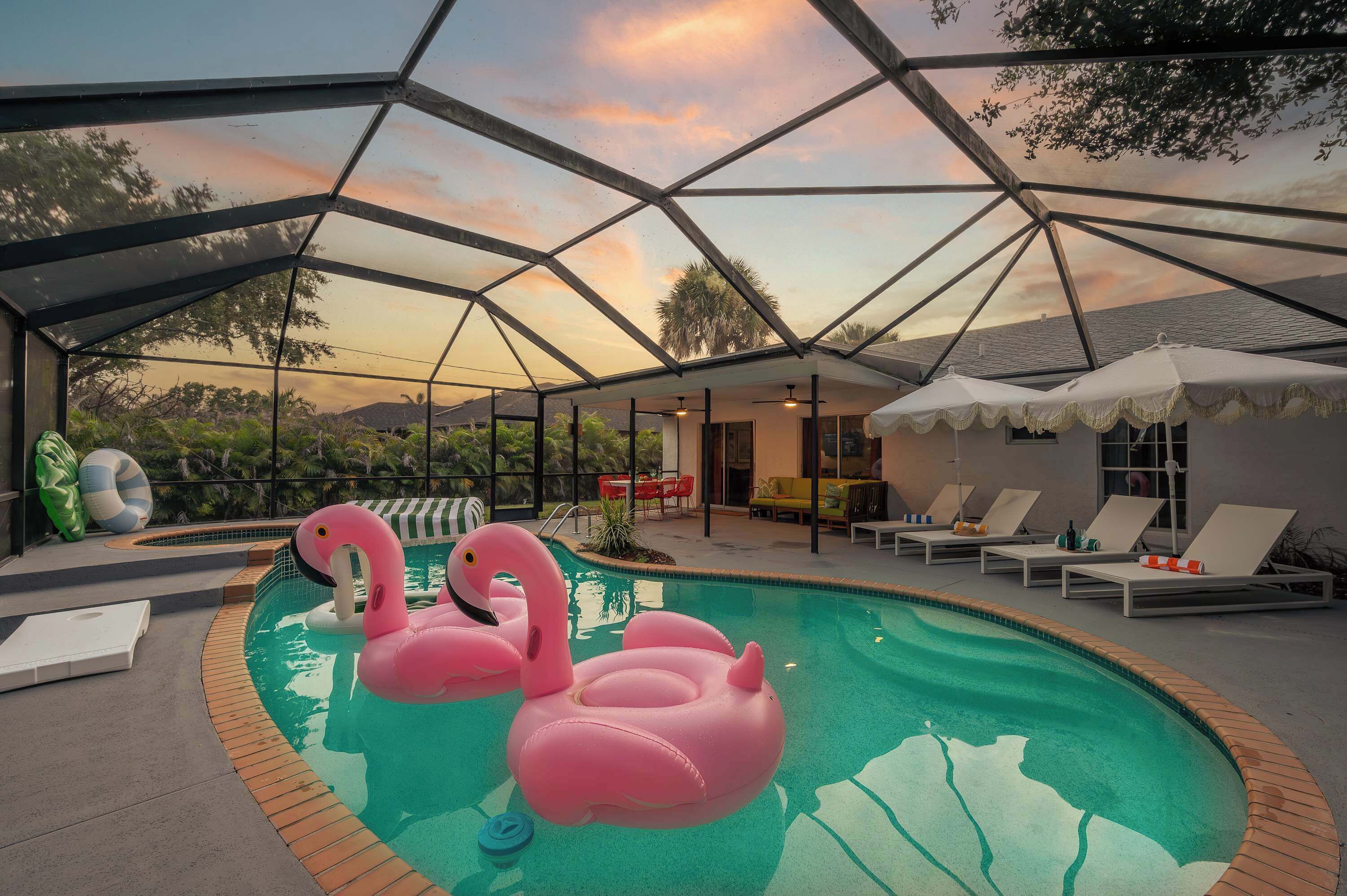 A swimming pool with inflatable flamingos, lounge chairs, and a covered area with a view of the sunset.