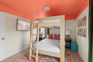 Habitació amb lliteres, un televisor penjat a la paret, una taula petita amb una làmpada i una maleta verda.