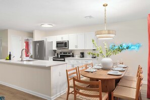 A modern kitchen with white cabinetry, stainless steel appliances, and a dining area with a round table and wicker chairs.