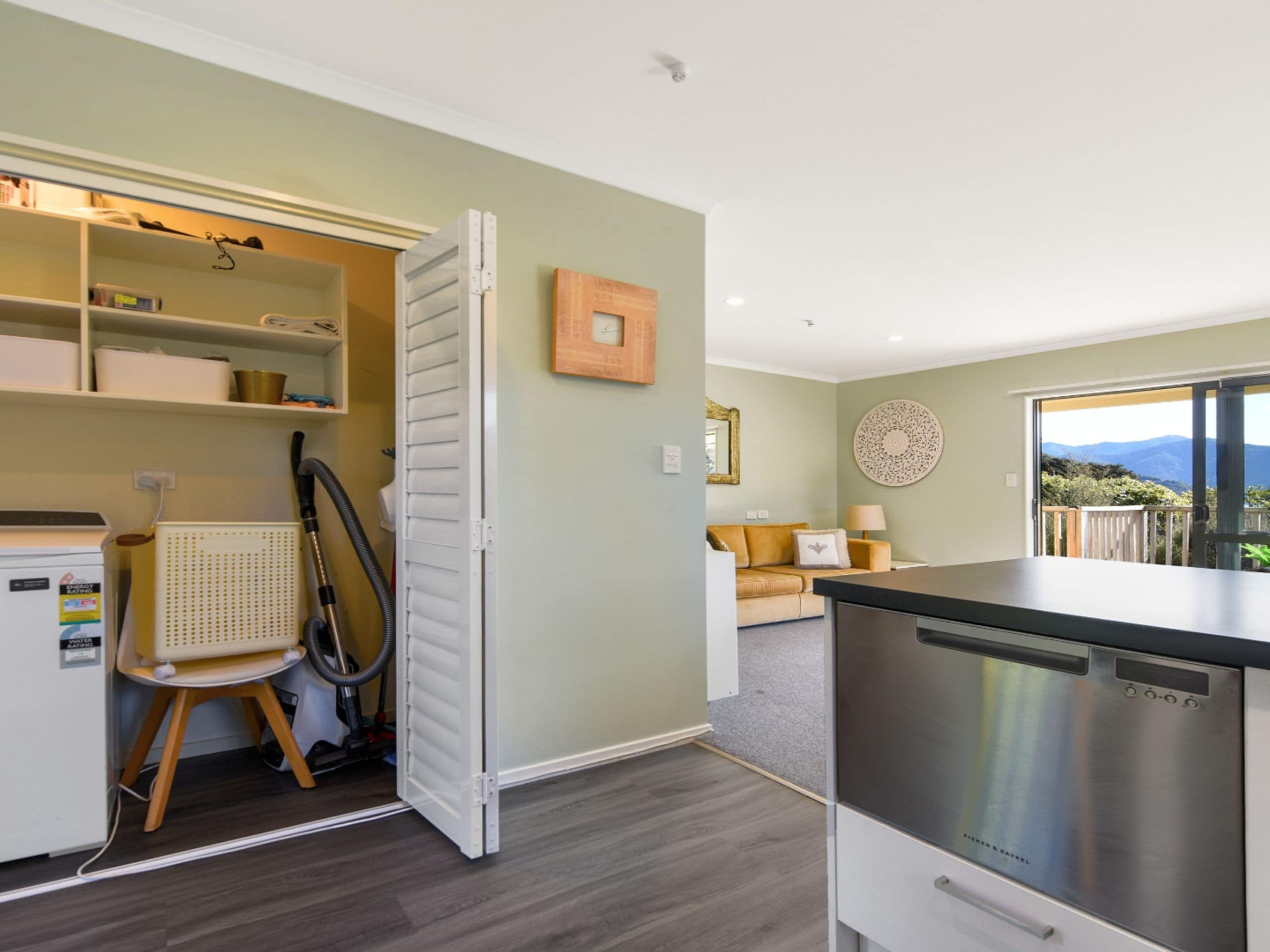 A compact kitchen with a dishwasher, a vacuum cleaner, and a dining area with a sofa and a table.