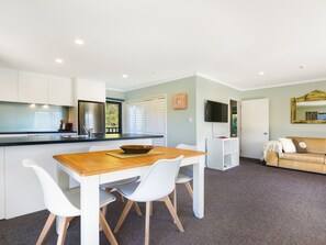 A modern kitchen with white cabinetry, a wooden dining table, and a TV mounted on the wall. - Tepuia Cottage - Havelock Holiday Retreat (Picton)